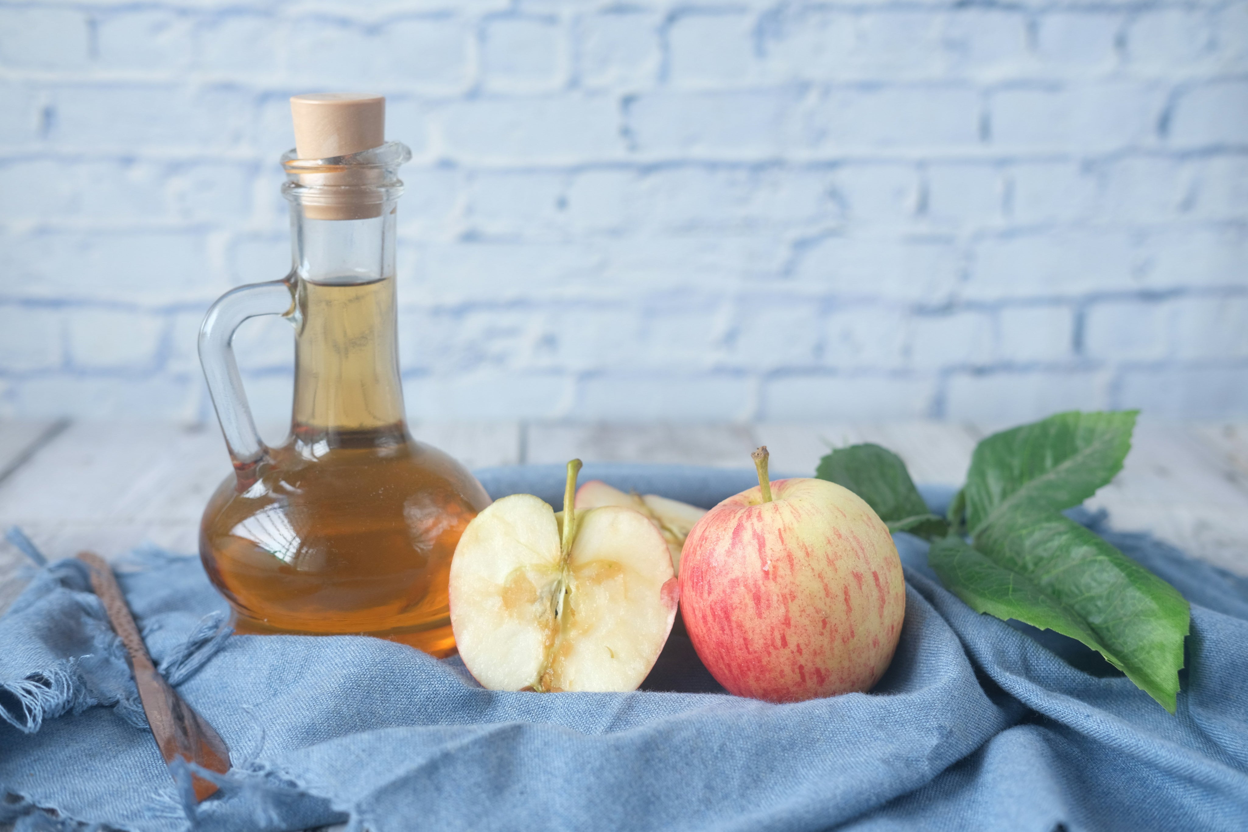 The image presents a rustic still life composition centered around apple cider vinegar and fresh apples. A clear glass bottle with a looped handle and a cork stopper dominates the left side of the frame, filled with a golden-hued liquid – presumably apple cider vinegar. The bottle is positioned on a slightly distressed, whitewashed wooden surface. To the right of the bottle, a partially sliced apple reveals its pale flesh and seeds, alongside a whole, red-striped apple. A few green apple leaves are tucked beside the whole apple, adding a touch of freshness and natural color. The entire arrangement rests upon a casually draped, textured blue denim cloth, which adds a soft, tactile element to the scene. The background is a whitewashed brick wall, providing a bright and slightly textured backdrop that doesn't distract from the main subjects. The lighting appears soft and natural, casting gentle shadows and highlighting the textures of the wood, fabric, and fruit. The overall aesthetic is clean, healthy, and evokes a sense of natural wellness, suggesting the benefits of apple cider vinegar and the goodness of fresh apples. The composition feels deliberately arranged yet relaxed, creating a visually appealing and inviting image.